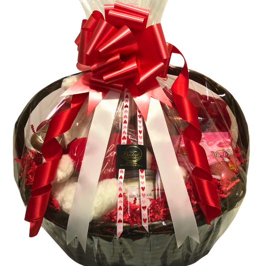 A romantic gift basket with a plush teddy bear, wine glass, chocolates, caramel popcorn, candle, and bath soak. Wrapped in clear cellophane with red bow, and red and pink heart ribbon.