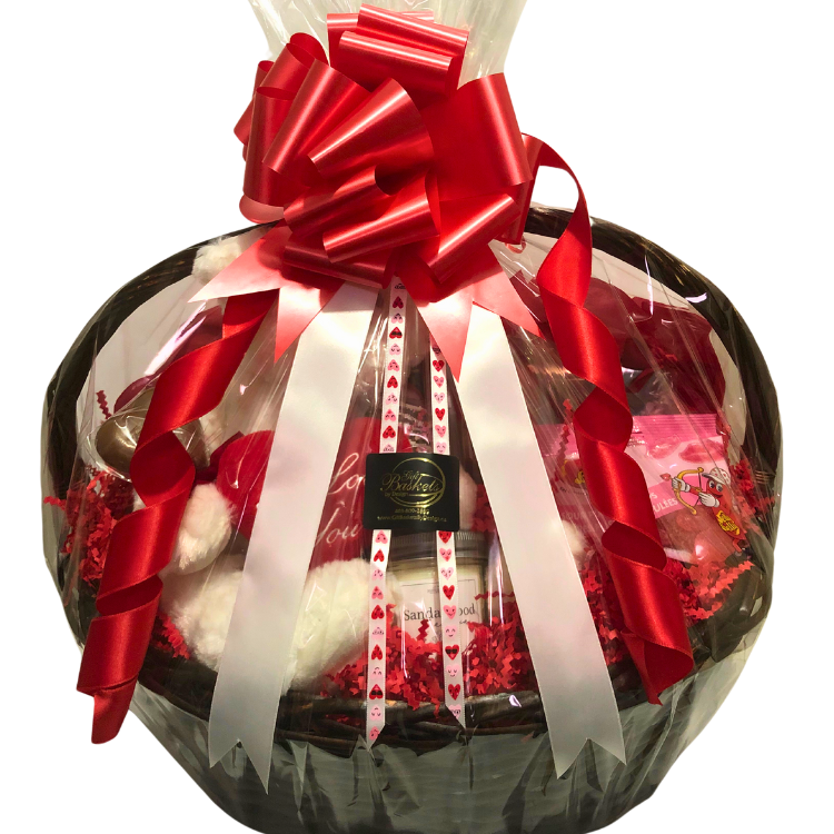 A romantic gift basket with a plush teddy bear, wine glass, chocolates, caramel popcorn, candle, and bath soak. Wrapped in clear cellophane with red bow, and red and pink heart ribbon.